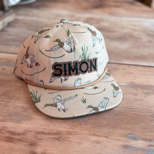May include: Tan baseball cap with a duck pattern and the name "SIMON" in dark brown letters. The cap features a brown brim and a rope detail. The duck pattern includes green and brown ducks and green reeds.