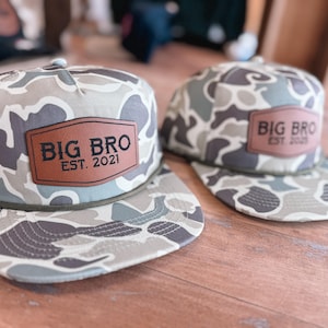 May include: Two camouflage baseball caps with brown leather patches that say "BIG BRO EST. 2021".