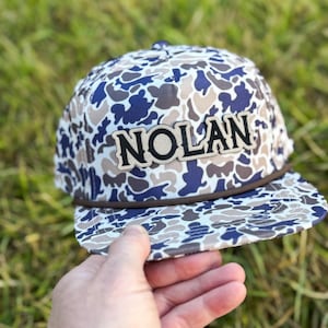 May include: A camouflage print baseball cap with the name "NOLAN" in bold, black letters. The cap features a brown brim and a matching brown rope detail. The background is blurred green grass.