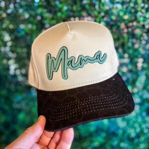 May include: A beige and black baseball cap with the word "Mama" in teal script across the front. The cap has a black brim and a structured design, suitable for casual wear.