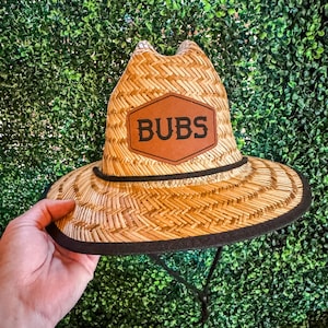 May include: A straw hat with a wide brim and a black trim. The hat features a brown leather patch with the word "BUBS" in white letters. It has a chin strap for a secure fit.