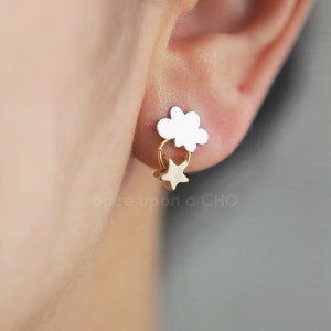 Puede incluir: Pendientes de oro con un dije de nube blanca y un dije de estrella colgante. Los pendientes se muestran en la oreja de una persona.