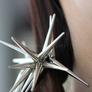 Peut inclure: Une pince à cheveux en forme d'étoile argentée avec de multiples pointes. La pince est dans les cheveux bruns foncés d'une femme.