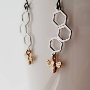 Puede incluir: Pendientes de panal de abeja plateados con un dije de abeja dorada que cuelga de cada pendiente.