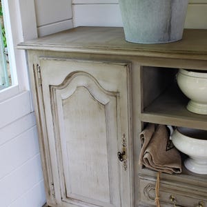 May include: A distressed, off-white cabinet with a curved door and brass fittings. The cabinet has open shelving with white bowls and a tan towel. A galvanised metal bucket with greenery sits on top, suggesting a farmhouse or cottage theme.