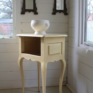 May include: A small, yellow painted side table with a shelf and ornate legs. The table is in a corner of a room with white walls and a white ceramic bowl on top.