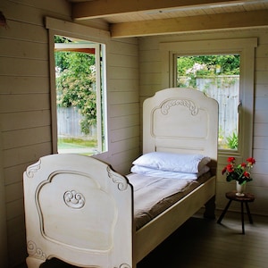 May include: A white wooden bed frame with a carved headboard and footboard. The bed is made with a white duvet and a brown blanket. The bed is in a room with wooden walls and a window with a view of a garden.
