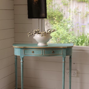 May include: A teal console table with an oval top, two drawers, and gold detailing. The table, with fluted legs, has a white ceramic bowl with branches on top. A framed portrait hangs on the wall. The table is approximately 100 cm long.