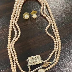 May include: A three-strand pearl necklace with a rectangular rhinestone clasp and matching pearl stud earrings. The necklace features a large pearl pendant and a gold filigree accent.