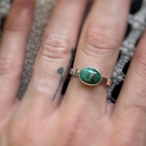 May include: A silver ring with an oval, green stone. The ring has a simple band and a bezel setting. The stone has a mottled appearance with shades of green and brown. The ring is shown on a hand.