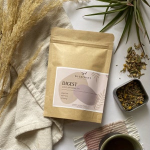 May include: A brown paper pouch with a label that reads "Wildcraft" and "Digest Loose Leaf Herbal Tea". The label also includes the words "digestive", "warming", and "relieving". The pouch is sitting on a white surface with a small tin of dried herbs and a cup of tea.