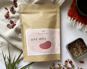 May include: A brown paper pouch with a white label that reads "Love Spell Loose Leaf Herbal Tea" with the words "warming, spicy, sensual" listed below. The pouch is sitting on a white surface with dried rose petals scattered around it.