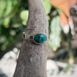 May include: A silver ring with an oval, teal-colored stone. The ring is set on a textured, gray surface, with a blurred green and orange background. The ring's band is thin and simple, highlighting the vibrant stone.