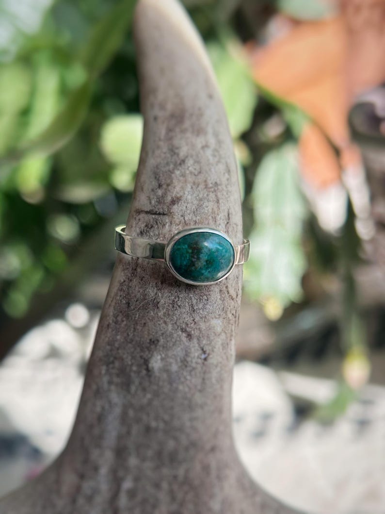 May include: A silver ring with an oval, teal-colored stone. The ring is set on a textured, gray surface, with a blurred green and orange background. The ring's band is thin and simple, highlighting the vibrant stone.