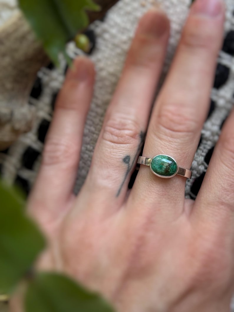 May include: A silver ring with an oval, green stone. The ring has a simple band and a bezel setting. The stone has a mottled appearance with shades of green and brown. The ring is shown on a hand.