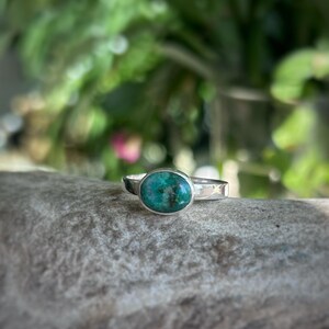 May include: A silver ring with an oval, turquoise-colored stone. The ring is set on a textured band. The background is blurred, with green foliage and a hint of pink flowers.