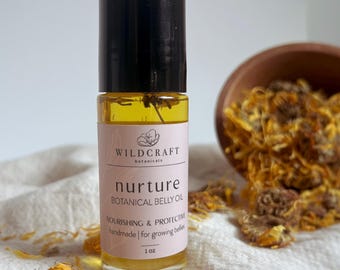 May include: A clear glass bottle of "nurture" botanical belly oil with a black cap. The label is pink and white with the text "Wildcraft Botanicals". The bottle is 1 oz. and is next to a small wooden bowl filled with dried orange flower petals.