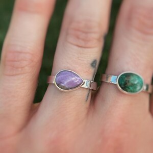 May include: Two rings with gemstone settings are displayed. One ring features a teardrop-shaped purple stone, while the other has an oval-shaped green stone. Both rings have a silver band.
