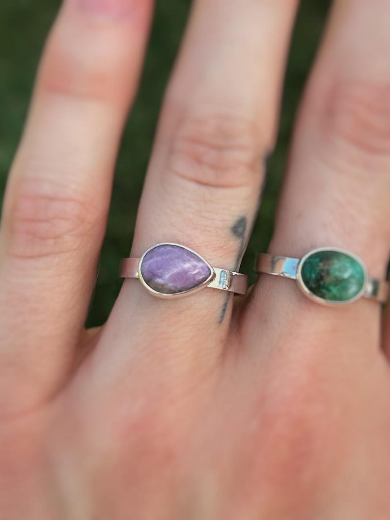 May include: Two rings with gemstone settings are displayed. One ring features a teardrop-shaped purple stone, while the other has an oval-shaped green stone. Both rings have a silver band.