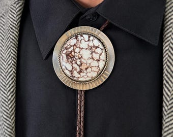 May include: A bolo tie featuring a circular metal setting with a sunburst-style edge and a central stone with a white and brown marbled pattern. The cord is made of braided dark brown leather, secured by a twisted metal rope border.