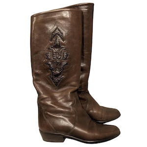 Puede incluir: Un par de botas altas de cuero marrón. Cada bota presenta un diseño decorativo en relieve en la caña superior. Las botas tienen un tacón bajo y una puntera ligeramente puntiaguda. El cuero tiene un aspecto desgastado y vintage.