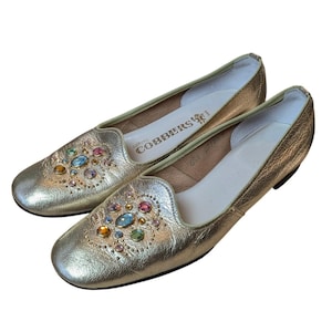 Può includere: Un paio di ballerine dorate in pelle con una decorazione di pietre preziose colorate sulla tomaia. Le scarpe hanno una punta arrotondata e un tacco basso. Il marchio "COBBERS" è visibile sulla soletta.