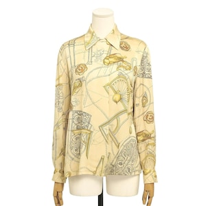 May include: A long-sleeved button-down shirt in a cream color, featuring a repeating pattern of gold and gray illustrations. The design includes ornate chairs, wheels, and the letter "E". The shirt has a classic collar and buttoned cuffs.