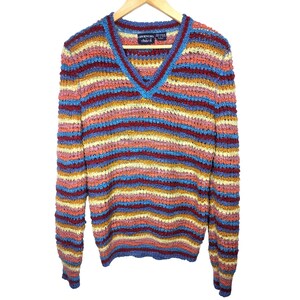 Pode incluir: Um suéter listrado multicolorido com decote em V. O suéter apresenta listras horizontais em tons de azul, vermelho, amarelo e rosa. A etiqueta diz "GIVENCHY Chara". O suéter está pendurado em um cabide de madeira.