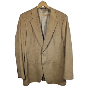 Könnte beinhalten: Ein beigefarbener Blazer mit zwei Knöpfen und zwei Vordertaschen, der an einem Holzbügel hängt. Der Blazer hat ein fallendes Revers und drei Knöpfe an jedem Ärmel. Das Innenfutter ist ein hellbraunes Stoffmuster. Der Blazer ist aus einem strukturierten Material.