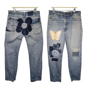 Può includere: Jeans in denim azzurro con una grande toppa a forma di fiore blu scuro sul retro. La parte anteriore presenta una toppa a forma di farfalla color crema, una toppa con faccina sorridente blu scuro e diverse toppe rettangolari.