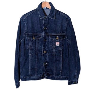 May include: A dark blue denim jacket featuring a classic collar, long sleeves, and a button-up front. It includes two chest pockets with buttoned flaps and a small rectangular white label with red text on the left pocket. The jacket has visible stitching.