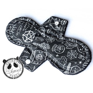 May include: A reusable cloth menstrual pad with a black background and white occult-themed illustrations. The pad features a snap closure and is decorated with pentagrams, spiderwebs, and other mystical symbols. The edges are lined with black fabric.