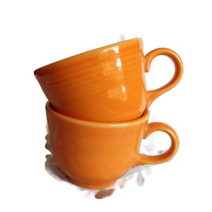 Peut inclure: Deux tasses en céramique orange empilées l'une sur l'autre. Les tasses ont une finition lisse et brillante et une forme arrondie.