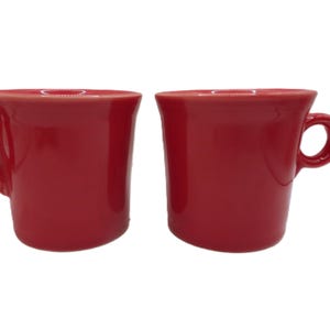 Może przedstawiać: Dwa pasujące do siebie jaskrawoczerwone kubki ceramiczne o gładkim, błyszczącym wykończeniu. Każdy kubek ma szeroki, rozszerzający się brzeg oraz okrągłe ucho przymocowane z boku. Kubki mają prosty, cylindryczny kształt z płaską podstawą.