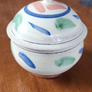 May include: A white ceramic lidded jar with a rounded shape. The lid and body are decorated with blue, green, and pink brushstrokes. A blue ring encircles the lid, and a small white knob tops it. The jar sits on a wooden surface.