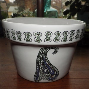 Puede incluir: Maceta de cerámica blanca con un diseño paisley verde, marrón y morado. La maceta tiene un borde decorativo alrededor de la parte superior.