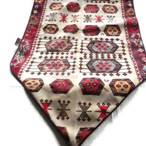 Puede incluir: Un camino de mesa blanco con un patrón geométrico rojo, marrón y azul. El camino de mesa tiene una borla de flecos en el extremo.