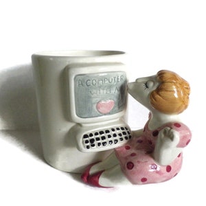 May include: A white ceramic mug shaped like a computer with a woman in a pink polka dot dress kissing the screen. The computer screen says "A Computer With A..." and has a pink heart on it.