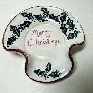 Può includere: Un piatto natalizio in ceramica bianca con bordo smerlato e forma unica. Il piatto è decorato con foglie di agrifoglio verde scuro e bacche rosse. Le parole "Merry Christmas" sono scritte in rosso al centro.