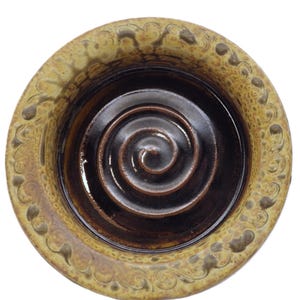May include: A ceramic bowl with a spiral design in the centre. The bowl has a dark brown interior and a light brown exterior with a decorative pattern around the rim. The bowl is round and sits on a white background.