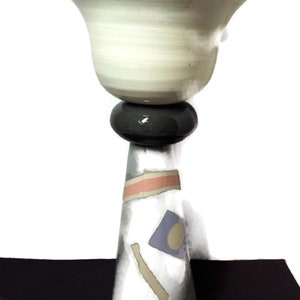 Puede incluir: Un macetero de cerámica blanca con un cuenco ancho y poco profundo en la parte superior de una base cónica alta. La base está decorada con formas abstractas en tonos rosa, azul y beige.