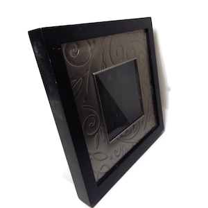 Puede incluir: Un marco de fotos cuadrado negro con un inserto decorativo plateado con un patrón floral en espiral. El marco tiene una pequeña abertura cuadrada en el centro para una foto. El marco está en ángulo, mostrando su profundidad.