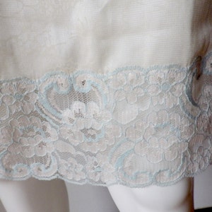 May include: Close-up of a cream-colored garment with a delicate floral pattern. The hem is trimmed with intricate white lace, accented with a light blue trim. The fabric appears soft and lightweight, suggesting lingerie or a similar item.