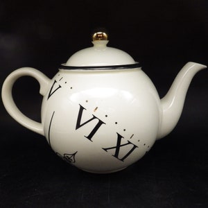 May include: White ceramic teapot with a black and gold clock face design. The teapot has a round body and a black handle. The clock face features Roman numerals and a gold knob on the lid.