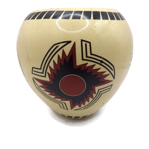 Può includere: Un vaso in ceramica color crema con un design geometrico nero e rosso. Il design presenta un sole stilizzato con raggi rossi e un centro nero.