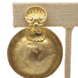 Pode incluir: Brincos dourados com um design circular. Cada brinco apresenta um elemento superior decorativo com um padrão semelhante a uma concha e uma base grande, plana e redonda. Os brincos são exibidos em um suporte de cor neutra.