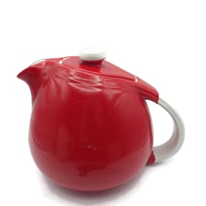 Op de afbeelding: Een glanzend rode keramische theepot met een witte handgreep en deksel. De theepot heeft een afgeronde body en een gebogen tuit. Het deksel is klein en rond, met een witte knop. De handgreep is gebogen.