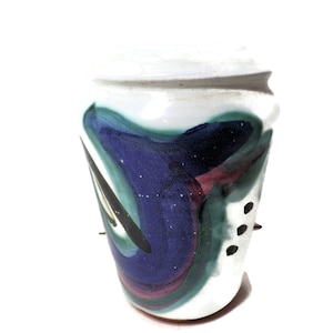 Może przedstawiać: Biały ceramiczny wazon z abstrakcyjnym wzorem w kolorze niebieskim, zielonym i fioletowym. Wazon ma zaokrąglony kształt i lekko rozszerzający się otwór.