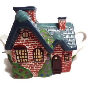 May include: A red brick house shaped teapot with a blue roof, green bushes, and a brown door. The teapot has a white spout and handle.