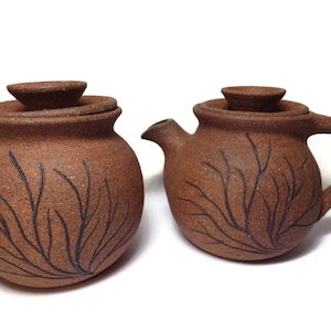 Puede incluir: Un juego de dos teteras de cerámica marrón con diseños de ramas de árboles negros. Las teteras tienen tapas y están hechas a mano.